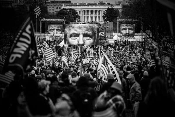 A black-and-white photo of people holding Trump flags and American flags and screens displaying Mr. Trump’s face in front of the White House. 